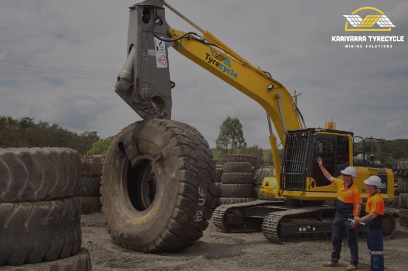 Excavator lifting large mining tyre ready for processing, workers standing in front